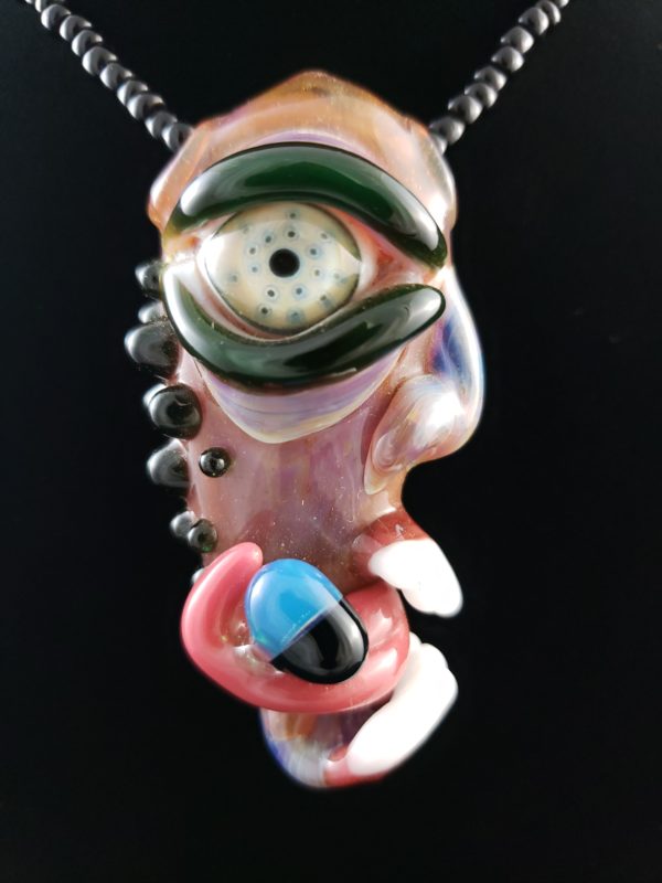 green eyed glass creature pendant with tongue holding a blue and black pill in mouth